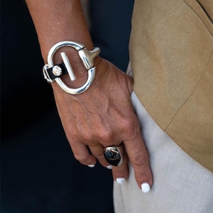 Puede incluir: Una pulsera plateada con un diseño grande y circular y una correa de cuero negro. La pulsera presenta una barra rectangular y un elemento decorativo. También es visible un anillo plateado a juego con una piedra oscura.