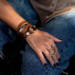 May include: A collection of gold and brown jewelry. A brown leather wrap bracelet with a gold buckle and a gold metal band. A gold ring with an abstract design is on the hand. The person is wearing blue jeans.