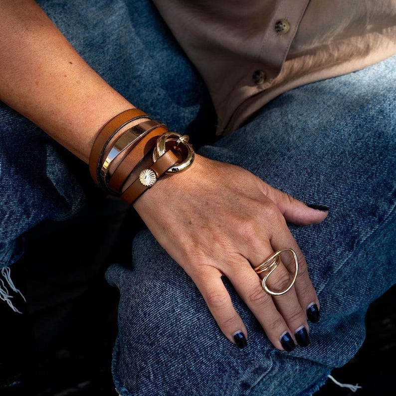 May include: A collection of gold and brown jewelry. A brown leather wrap bracelet with a gold buckle and a gold metal band. A gold ring with an abstract design is on the hand. The person is wearing blue jeans.