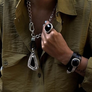 May include: A silver necklace with a black pendant, a matching ring, and a black leather bracelet with a silver accent. The jewellery features a bold, geometric design with black stone inlays. The person is wearing an olive green shirt.