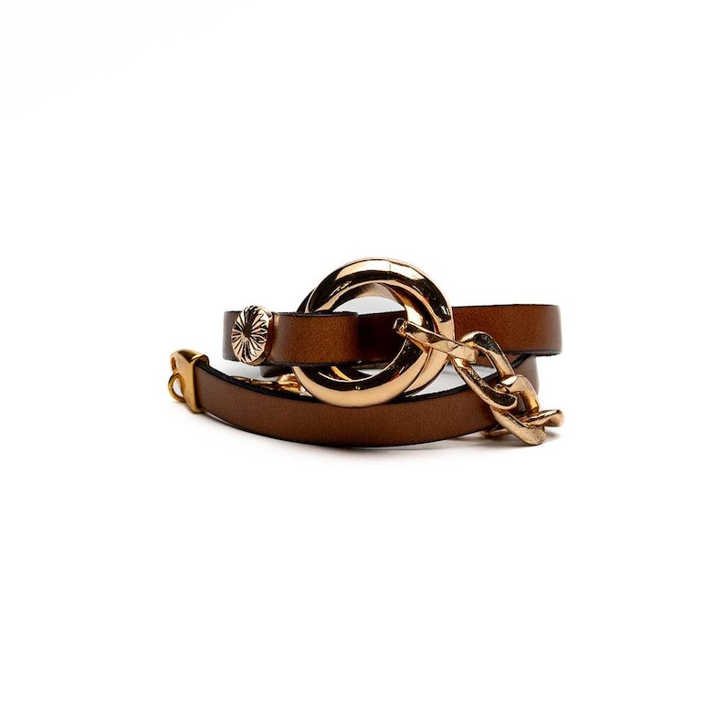 May include: A brown leather bracelet with a gold-colored chain and a large gold-colored circular clasp. The bracelet has a small gold-colored decorative element and a gold-colored buckle.