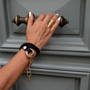 Puede incluir: Una pulsera de cuero negro con un anillo y una cadena dorados. Un anillo dorado se lleva en la mano, que sostiene una manija de puerta de bronce. El fondo es una puerta gris.
