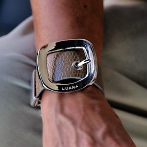 May include: A wide bracelet featuring a textured, tan-colored band with a snakeskin-like pattern. The centerpiece is a large, polished silver-tone metal buckle with a square shape and rounded corners. The word LUANA is engraved on the buckle.