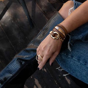 May include: A gold ring with a leaf design and a multi-strand brown leather bracelet with gold accents. The bracelet features a large gold clasp. The person is wearing blue jeans and black boots.