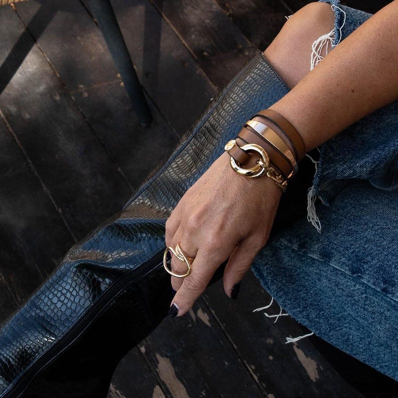 May include: A gold ring with a leaf design and a multi-strand brown leather bracelet with gold accents. The bracelet features a large gold clasp. The person is wearing blue jeans and black boots.