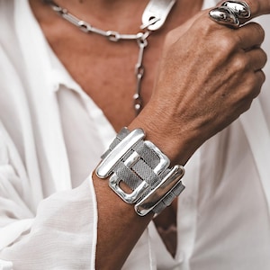 Può includere: Un ampio bracciale rigido color argento caratterizzato da un pannello centrale in maglia metallica scura incorniciato da barre rettangolari lisce e lucide color argento. Il bracciale è indossato con una collana a catena e un grande anello liscio.