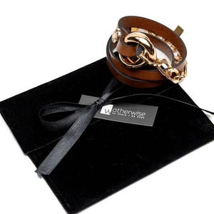 May include: A brown leather wrap bracelet with a gold-colored chain and clasp. The bracelet is displayed on a black velvet pouch with a black ribbon and a tag that reads "Wotherwise be sharp / be edgy."