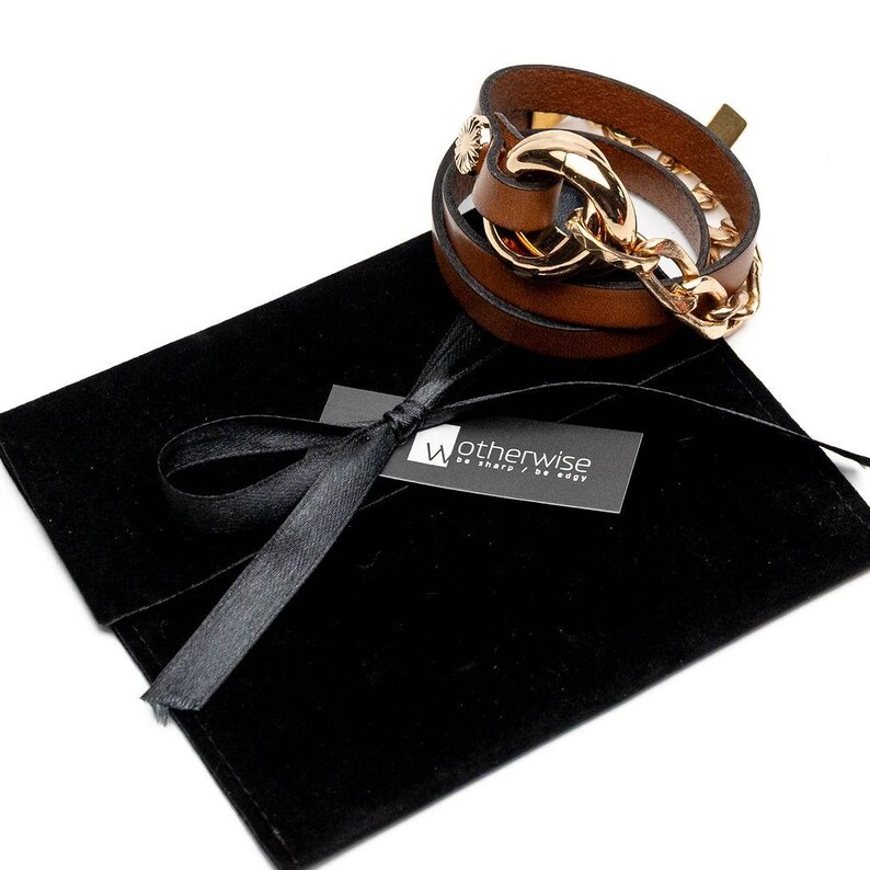 May include: A brown leather wrap bracelet with a gold-colored chain and clasp. The bracelet is displayed on a black velvet pouch with a black ribbon and a tag that reads "Wotherwise be sharp / be edgy."