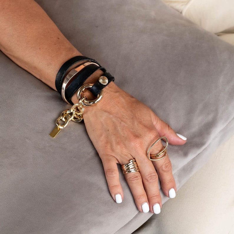 Puede incluir: Una pulsera de cuero negro con detalles dorados y un dije de cadena dorada. La pulsera se combina con anillos dorados, incluido un anillo ovalado grande y un anillo de varias bandas. Las joyas se muestran en una mano.