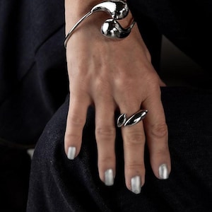 Op de afbeelding: Een zilverkleurige armband- en ringenset. De armband heeft twee afgeronde, druppelvormige elementen. De ring heeft een vergelijkbaar ontwerp met een enkele druppelvorm. De sieraden worden gedragen op een hand met zilveren nagellak.