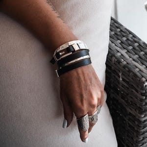 Op de afbeelding: Een zilveren en zwarte leren wikkelarmband met een gespsluiting wordt om een pols gedragen. Verschillende zilveren ringen met sierlijke ontwerpen sieren de vingers. De persoon draagt een witte jurk.