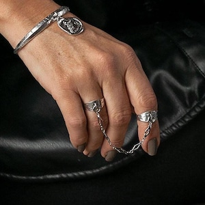 Può includere: Un bracciale d'argento con un ciondolo e due anelli d'argento collegati da una catena. Gli anelli sono indossati su dita adiacenti. Il bracciale ha un design strutturato. Il ciondolo è rotondo con un design dettagliato.