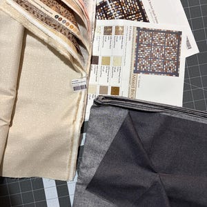 Könnte beinhalten: Ein Quilt-Set mit einem Stapel gefalteter Stoffe in Creme-, Braun- und Beigetönen sowie einem dunkelgrauen Stoffstück. Enthalten sind gedruckte Anleitungsblätter mit dem Titel Mixed Bricks Quilt Pattern, die Stoffmuster und Schritte zeigen.