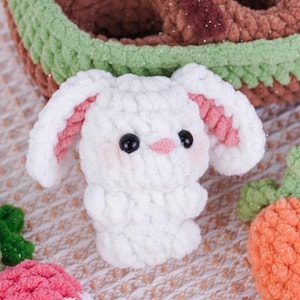 May include: A white crochet bunny with pink inner ears, black eyes, and a pink nose. The bunny has rosy cheeks and is part of a collection of handmade crochet toys. The bunny is shown with a crochet carrot and a basket.