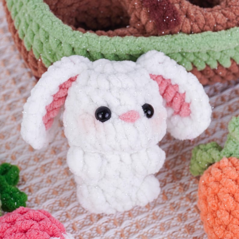May include: A white crochet bunny with pink inner ears, black eyes, and a pink nose. The bunny has rosy cheeks and is part of a collection of handmade crochet toys. The bunny is shown with a crochet carrot and a basket.