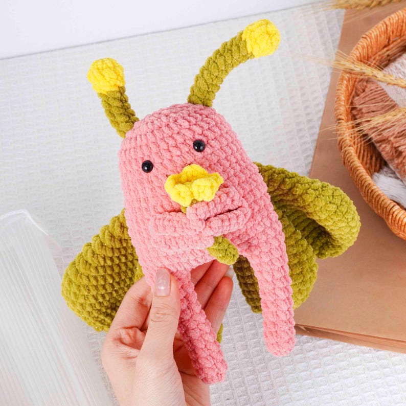 Puede incluir: Un juguete de crochet hecho a mano, rosa y verde, con detalles amarillos. El juguete tiene ojos negros y est&aacute; siendo sostenido. El bicho tiene un aspecto texturizado y afelpado, con un dise&ntilde;o caprichoso.