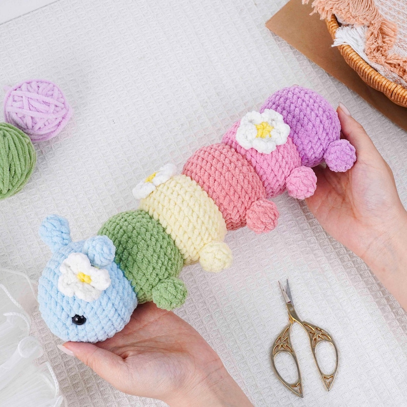 Puede incluir: Un juguete de oruga de ganchillo hecho a mano, en tonos pastel de azul, verde, amarillo, rosa y morado, cada segmento con una flor blanca. El juguete se sostiene con dos manos, con hilo y tijeras en el fondo.