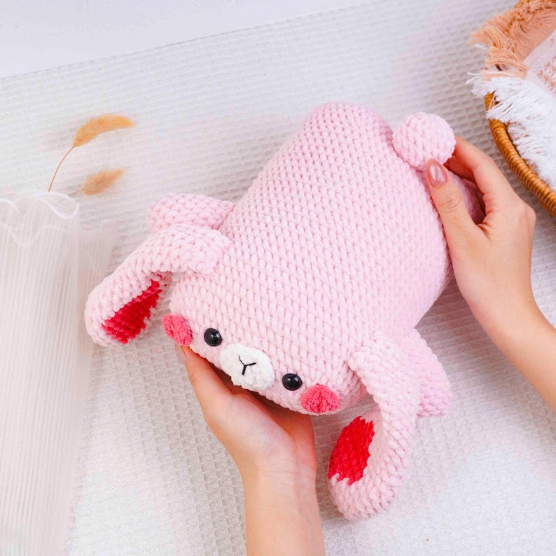 Op de afbeelding: Een handgemaakt roze gehaakt konijn knuffel met rode accenten op de oren en voeten. Het konijn heeft zwarte ogen en roze wangen. Het speelgoed is zacht en knuffelbaar.
