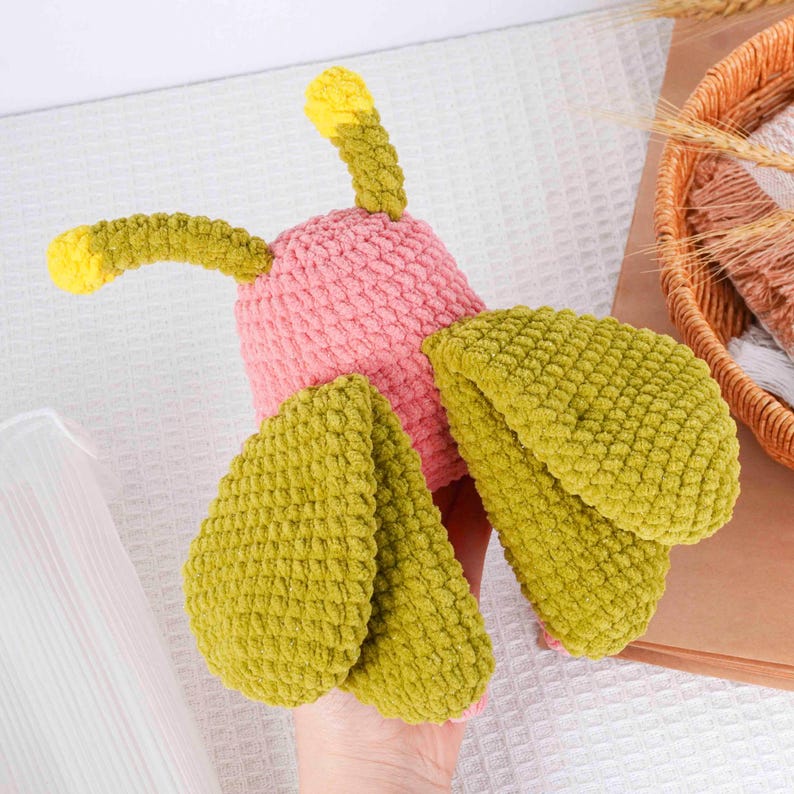 Puede incluir: Un juguete de ganchillo hecho a mano con forma de insecto colorido. El juguete presenta un cuerpo rosa, alas verdes y antenas verdes y amarillas. El hilo suave y texturizado le da al juguete un aspecto caprichoso.
