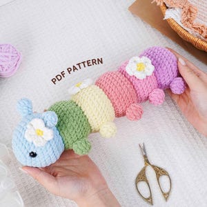 May include: A colorful, crocheted caterpillar toy in shades of blue, green, yellow, pink, and purple, adorned with white flower accents. The text "PDF PATTERN" is visible. A pair of gold scissors and yarn balls are also present.