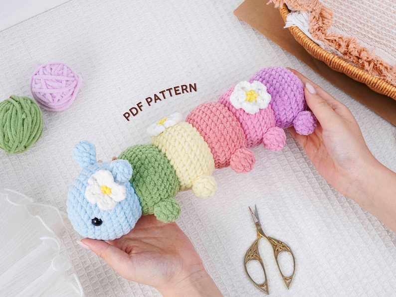 Puede incluir: Un juguete de oruga de ganchillo multicolor en tonos azul, verde, amarillo, rosa y morado, adornado con flores blancas. El texto "PDF PATTERN" es visible. Tambi&eacute;n hay unas tijeras doradas y ovillos de lana.