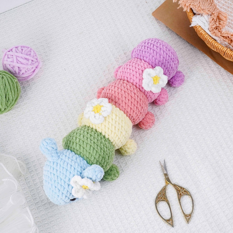 Puede incluir: Un juguete de oruga de ganchillo hecho a mano en colores pastel. La oruga est&aacute; hecha de hilo suave en tonos de azul, verde, amarillo, rosa y morado. Cada segmento tiene una flor blanca con un centro amarillo. Unas tijeras doradas y ovillos de lana tambi&eacute;n est&aacute;n en la imagen.