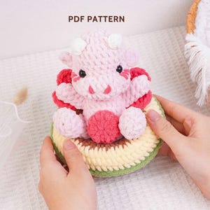 May include: A handmade crochet cow in shades of pink and red, with white horns and black eyes, sitting on a crocheted avocado. The text "PDF PATTERN" is visible at the top. The cow is held by hands.