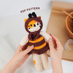 Puede incluir: Una muñeca de ganchillo hecha a mano con un disfraz de gato y un vestido a rayas de abeja. La muñeca tiene un vestido a rayas marrón y amarillo, una cara de gato y una capucha con antenas. El texto "PDF PATTERN" es visible.