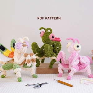 May include: Three crocheted mantis figures in cream with green and yellow wings, solid green with pink eyes, and solid pink with white accents. The text PDF PATTERN is centred above the figures. A pair of scissors and a crochet hook are below.