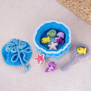 May include: A crocheted ocean-themed playset featuring a blue bowl with sea creatures, a drawstring bag, and a magnifying glass. The bowl contains a yellow fish, a starfish, and purple jellyfish. The magnifying glass has a yellow fish.