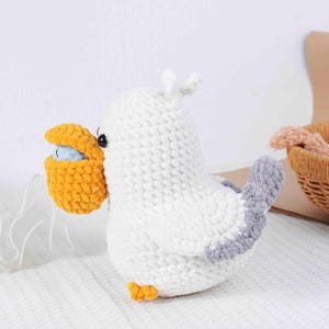 May include: A handmade crochet seagull toy in white, gray, and yellow. The seagull holds a yellow basket with a blue ball inside. The toy has black eyes and small yellow feet. The seagull's tail is gray.