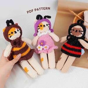 Puede incluir: Tres muñecas de gato hechas a mano con ganchillo, cada una con un atuendo diferente. Una está vestida de abeja en marrón y amarillo, otra con un vestido rosa con detalles de flores y la tercera con un atuendo negro con rayas rojas. Las muñecas tienen cuerpos blancos y caras de gato.