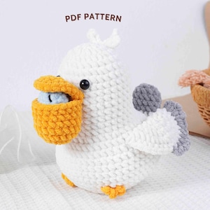以下が含まれることがあります： 黄色いポーチと灰色の翼が付いた、手作りの白いかぎ針編みの鳥。鳥は黒い目と黄色の足を持っています。「PDF PATTERN」という文字が上部にあります。このソフトスカルプチャーは、ユニークな室内装飾品です。