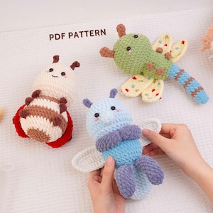 May include: Three crocheted plush toys featuring a PDF PATTERN label. One is a cream and brown striped insect, one is a green dragonfly with yellow wings, and one is a light blue and purple insect with white wings, held by hands.