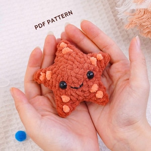 May include: A small, five-pointed starfish crochet plushie made of textured, burnt orange yarn with lighter orange spots. The starfish features two round, black plastic safety eyes and a stitched black mouth. The text PDF PATTERN is visible.