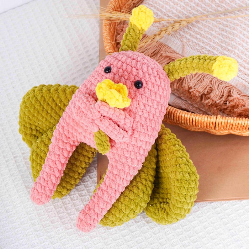Puede incluir: Un juguete de p&aacute;jaro de ganchillo hecho a mano en rosa, amarillo y verde. El p&aacute;jaro tiene un cuerpo texturizado, ojos negros y alas extendidas. El juguete est&aacute; hecho de hilo suave y est&aacute; dise&ntilde;ado para ni&ntilde;os.