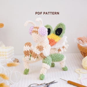 Puede incluir: Una figura de mantis religiosa tejida a ganchillo con hilo crema, marrón y verde claro con detalles en lavanda en la cabeza. El texto PDF PATTERN es visible sobre la figura. La criatura tiene grandes ojos verdes y negros y extremidades.