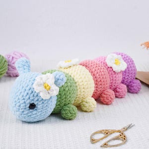 Puede incluir: Una fila de juguetes de oruga de ganchillo de colores pastel: azul, verde, amarillo, rosa y morado. Cada oruga tiene una flor blanca con centro amarillo y peque&ntilde;os ojos negros. Los juguetes est&aacute;n hechos a mano y colocados sobre una superficie blanca.