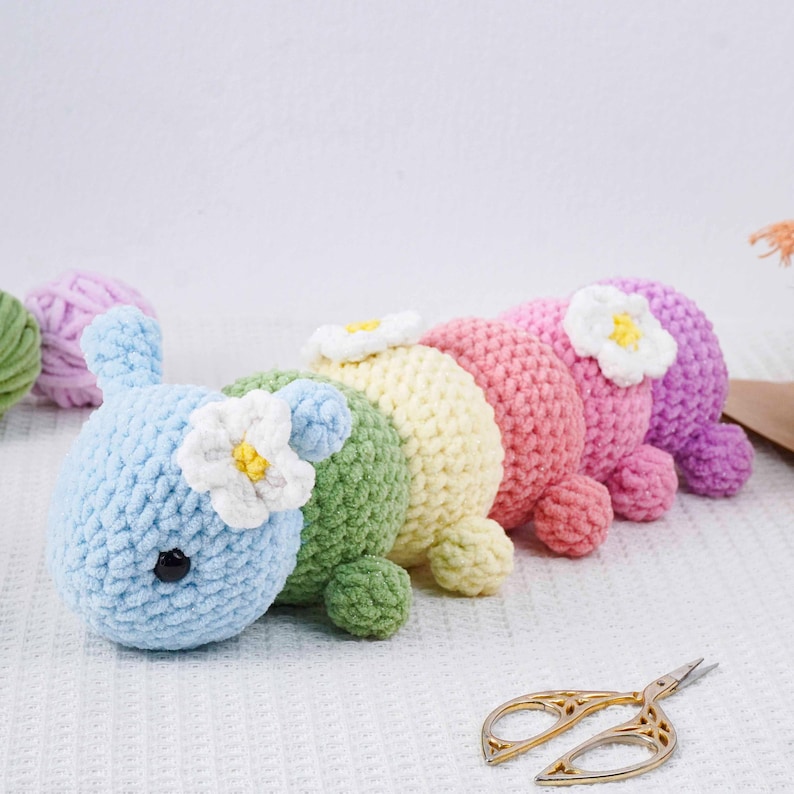 Puede incluir: Una fila de juguetes de oruga de ganchillo de colores pastel: azul, verde, amarillo, rosa y morado. Cada oruga tiene una flor blanca con centro amarillo y peque&ntilde;os ojos negros. Los juguetes est&aacute;n hechos a mano y colocados sobre una superficie blanca.