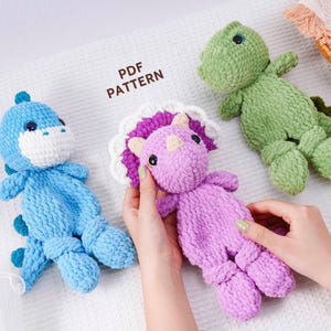 Puede incluir: Tres peluches de dinosaurios de ganchillo hechos a mano en azul, verde y morado. Una persona sostiene el dinosaurio morado. El texto "PDF PATTERN" es visible en la imagen. Estos juguetes blandos son perfectos para niños.