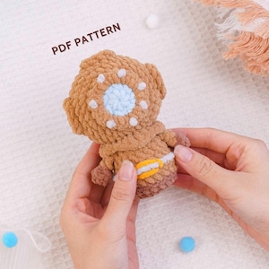 Pode incluir: Um brinquedo de pelúcia em crochê castanho com formato de capacete de mergulhador, apresentando uma janela circular azul-clara, detalhes brancos e um cinto amarelo. O texto PDF PATTERN está impresso em preto acima da figura sobre uma superfície.