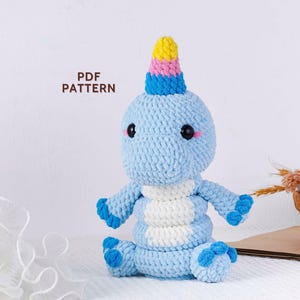 May include: A light blue crocheted dinosaur plushie with a white belly and a colourful horn. The dinosaur has black eyes and pink cheeks. The text "PDF PATTERN" is in the upper left corner. The plushie is sitting on a white surface.