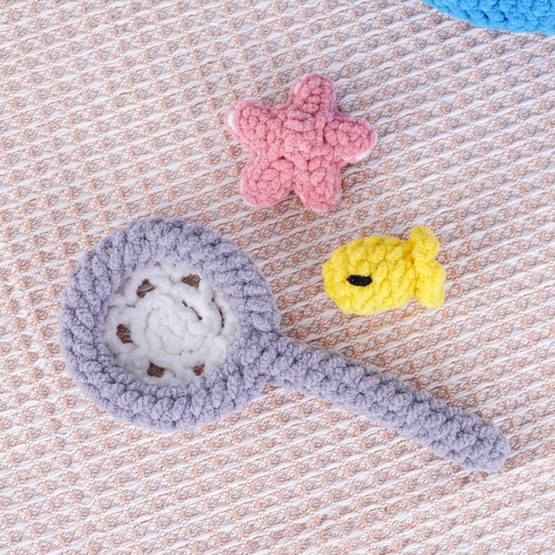 May include: A collection of handmade crochet toys, including a lavender bubble wand, a pink starfish, and a yellow fish with a black eye. The toys are made of soft, textured yarn and are arranged on a textured, beige surface.