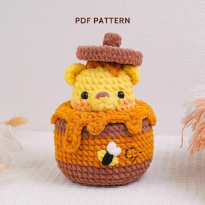 May include: A crochet amigurumi bear emerging from a brown and orange honey pot with a lid. The bear is yellow with black eyes and pink cheeks. The pot features a textured honey drip design and a small bee appliqué. Text reads PDF PATTERN.