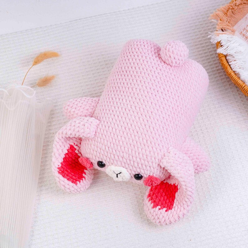 Op de afbeelding: Een lichtroze gebreid konijn knuffel met rode accenten op de oren. Het konijn heeft zwarte ogen en een witte snuit. Het speelgoed ligt op een wit oppervlak.