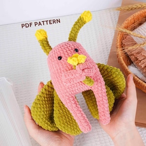 Puede incluir: Un peluche de ganchillo hecho a mano con forma de bicho caprichoso. El peluche es rosa con alas y antenas verdes y detalles amarillos. El texto "PDF PATTERN" es visible en el fondo.