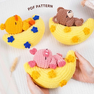 May include: Three crocheted baby toys: a yellow duck, a brown bear, and a pink pig, each resting on a yellow crescent moon. The moons have orange and blue or yellow accents. The text "PDF PATTERN" is visible.