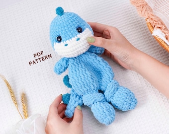 Pode incluir: Um brinquedo de dinossauro de crochê azul feito à mão com focinho e barriga brancos. O dinossauro tem olhos pretos pequenos e um pequeno espinho azul na cabeça. O texto "PDF PATTERN" é visível.