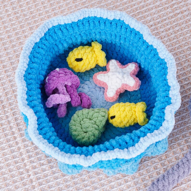 May include: A handmade, round, blue crochet basket filled with sea creatures. The basket has a wavy edge and contains a yellow fish, a purple jellyfish, a white starfish with pink trim, a green shell, and another yellow fish.