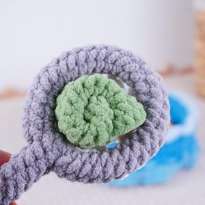May include: A handmade crochet toy in a whimsical design. The toy features a light purple ring with a green center, creating a soft, textured appearance. The plush material suggests a focus on comfort and tactile appeal, ideal for children's play.